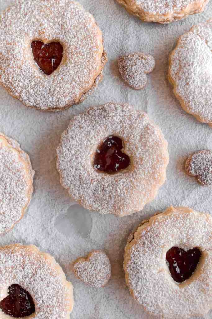 Shortbread linzer cookies on a parchment paper with heart shaped cutots and filled with raspberry jam