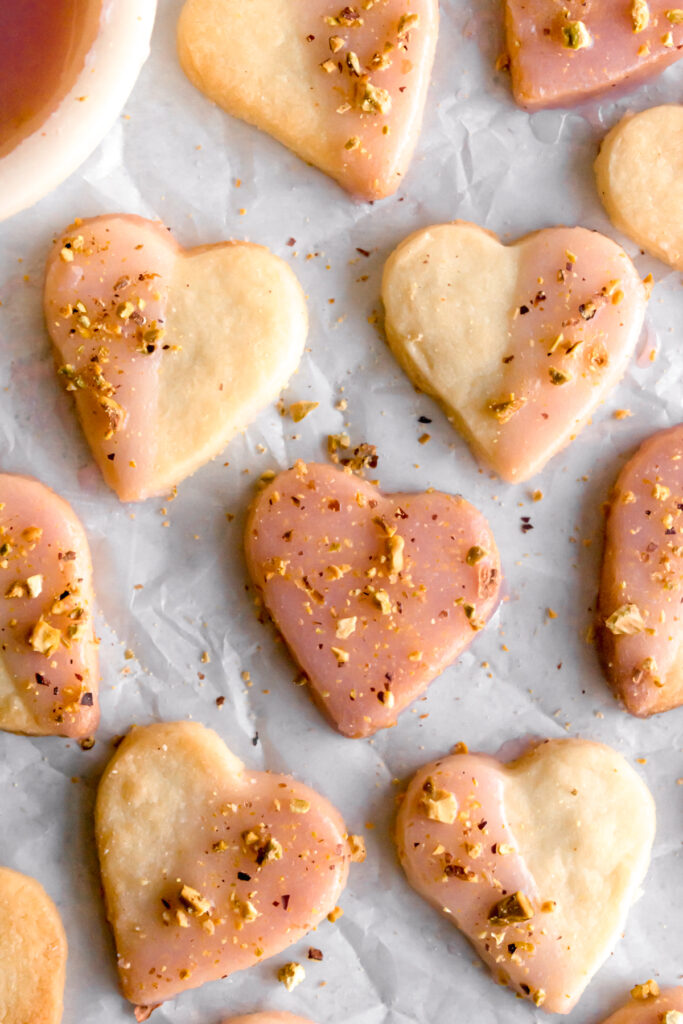 heart shaped shortbread cardamom cookies dipped in with rose flavoured glaze and topped with crushed pistachios on a parchment paper